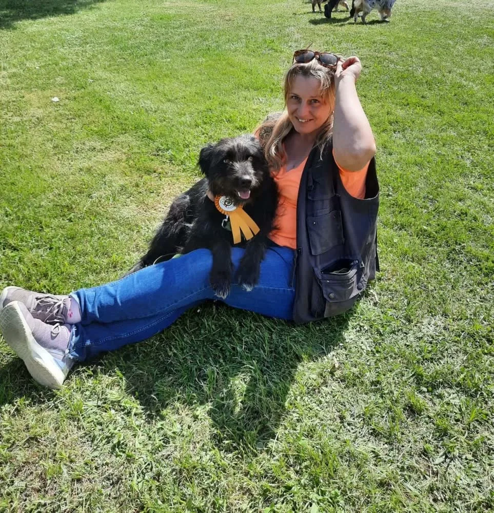 Frau sitzt lachend im Gras mit einem schwarzen Hund, der eine Siegerschleife trägt